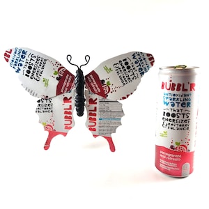 May include: A butterfly made from two aluminum cans of Bubbl'r Antioxidant Sparkling Water. The cans are decorated with red, white, and blue graphics and the text "That Boosts Energizes & Restores Balance". The butterfly is sitting on a white surface.
