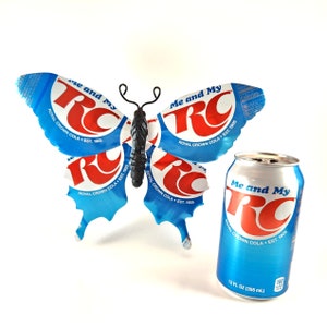 May include: A blue and red butterfly made from recycled Royal Crown Cola cans. The butterfly is next to a 12-ounce can of Royal Crown Cola.
