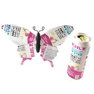 May include: A white can of Bubbl'r antioxidant sparkling water with a pitaya berry nectar flavor, next to a paper butterfly. The butterfly is made from the can's packaging, with the same text and graphics.