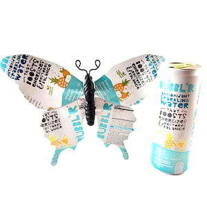 May include: A butterfly sculpture made from a beverage can, with a black body and wire antennae. The wings are made from the can's label, featuring text and a pineapple design. A matching can is next to it.
