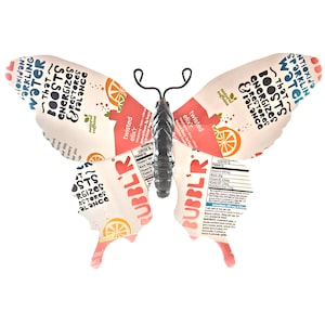 May include: A butterfly-shaped wall art made from recycled aluminum cans. The cans are from a sparkling water brand called "Bubbl'r" and feature the text "Twisted Elix'r" and "Sparkling Water That Boosts, Energizes & Restores Balance".