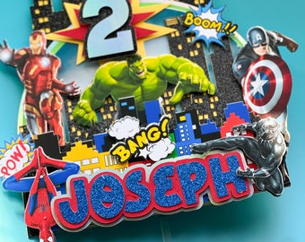 May include: A superhero cake topper with Iron Man, Hulk, Captain America, Black Panther, and Spider-Man. The number "2" is displayed, along with the name "Joseph" in blue glitter. Comic book sound effects and a cityscape backdrop complete the design.