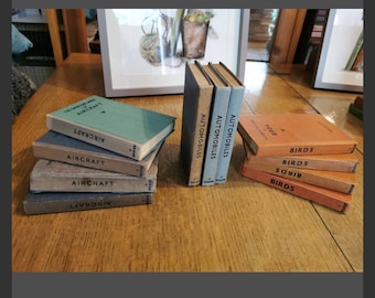 May include: A collection of vintage books with titles including "Aircraft", "Automobiles", and "Birds". The books have aged covers in various colors such as green, blue, and orange. The books are arranged on a wooden surface.