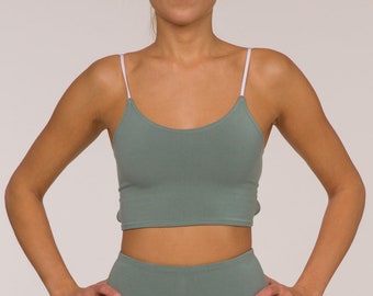Pode incluir: Conjunto desportivo verde sálvia, incluindo um top curto com alças brancas e calções de ciclista de cintura alta. A modelo usa ténis brancos. O conjunto é feito de um material macio e elástico, perfeito para treinos ou uso casual.