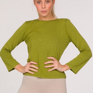 Puede incluir: Una persona con una camiseta verde oliva de manga larga y unos pantalones cortos de ciclista beiges. La camiseta tiene cuello redondo y los pantalones cortos llegan a la rodilla. El conjunto es sencillo y adecuado para actividades deportivas o informales.