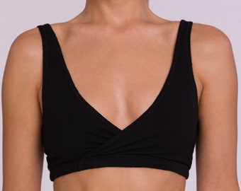Pode incluir: Top preto curto com design de decote em V. O top tem uma frente cruzada e alças largas. O tecido parece ser um material macio de cor sólida. A imagem mostra o top combinado com calças pretas a condizer.