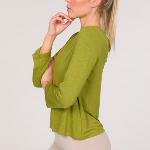 Puede incluir: Una mujer con una camiseta verde oliva de manga larga y leggings beige. La camiseta tiene cuello redondo y mangas acampanadas. Una goma blanca sujeta su pelo rubio en una cola de caballo.