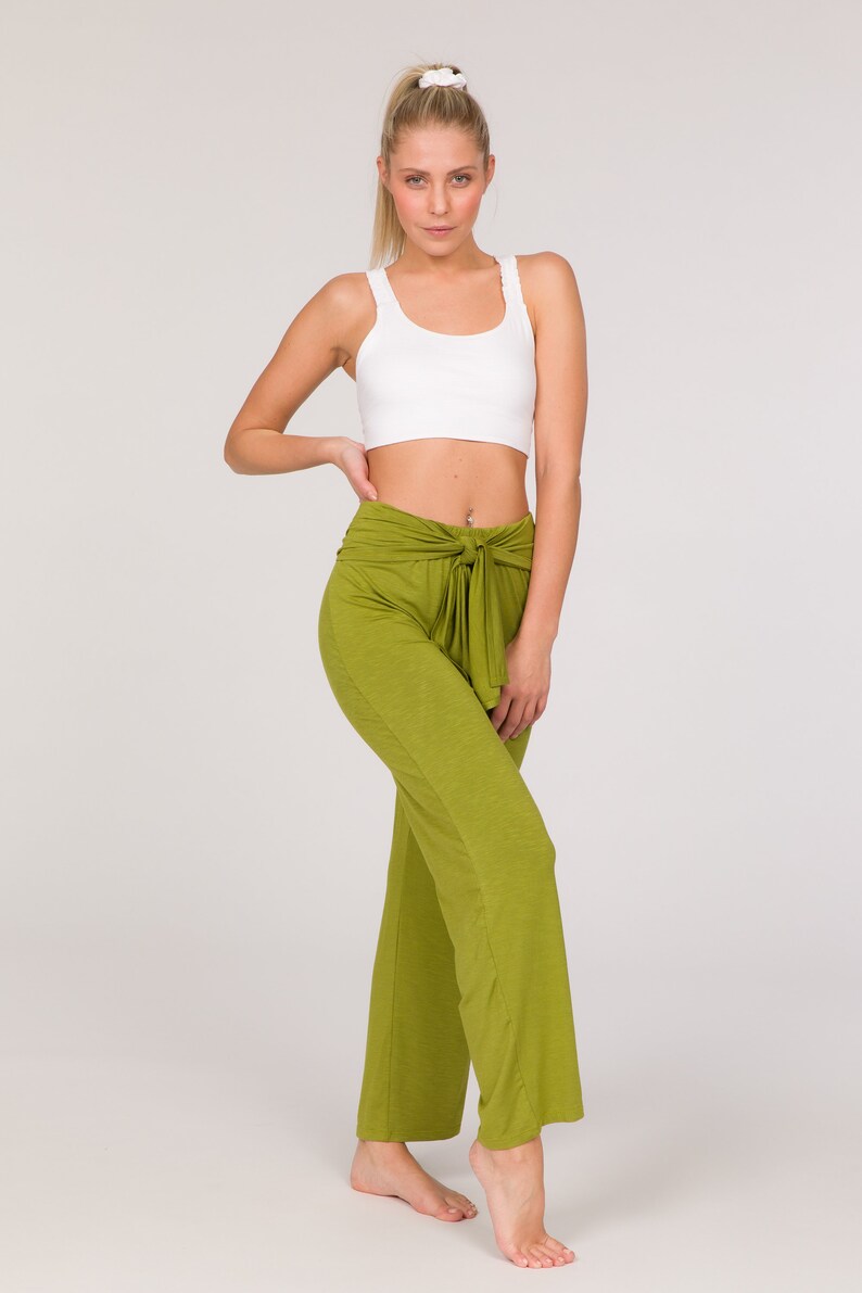 May include: A woman wearing a white sports bra and olive green wide-leg trousers with a tie waist. The trousers are a solid colour and have a relaxed fit. The model is standing barefoot against a white background.