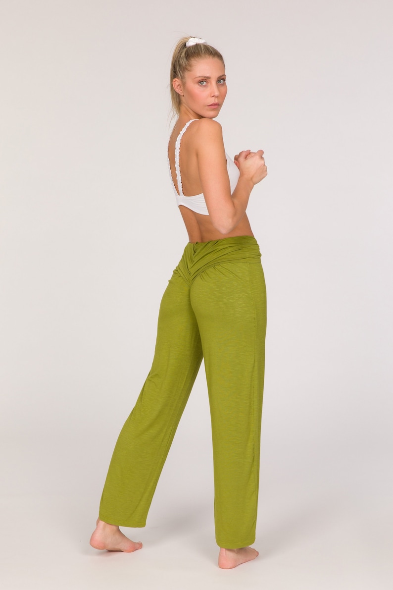 May include: A woman in a white sports bra and olive green yoga trousers. The trousers feature a ruched waist and a loose fit. The woman is turned away from the camera, looking over her shoulder.