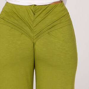 May include: Close-up of olive green trousers with a gathered waist detail. The trousers have a wide leg design and are made of a textured fabric. A white long-sleeved top is visible above the trousers.