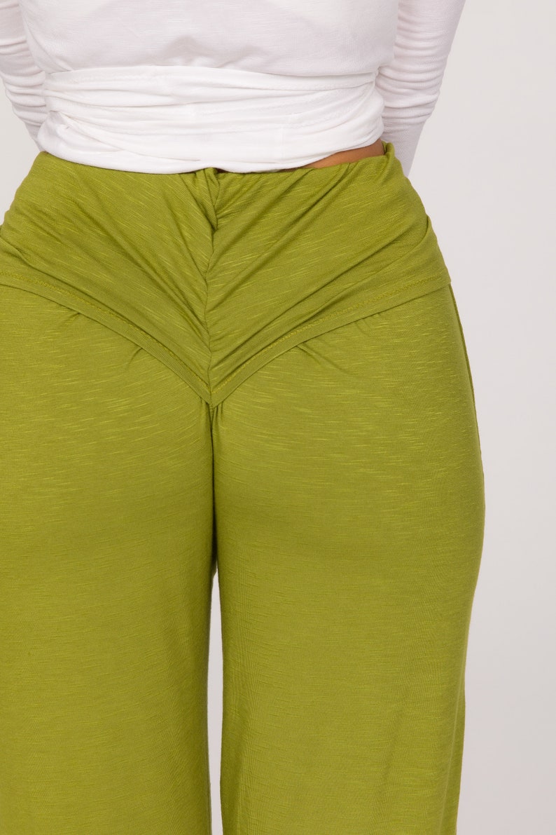 May include: Close-up of olive green trousers with a gathered waist detail. The trousers have a wide leg design and are made of a textured fabric. A white long-sleeved top is visible above the trousers.