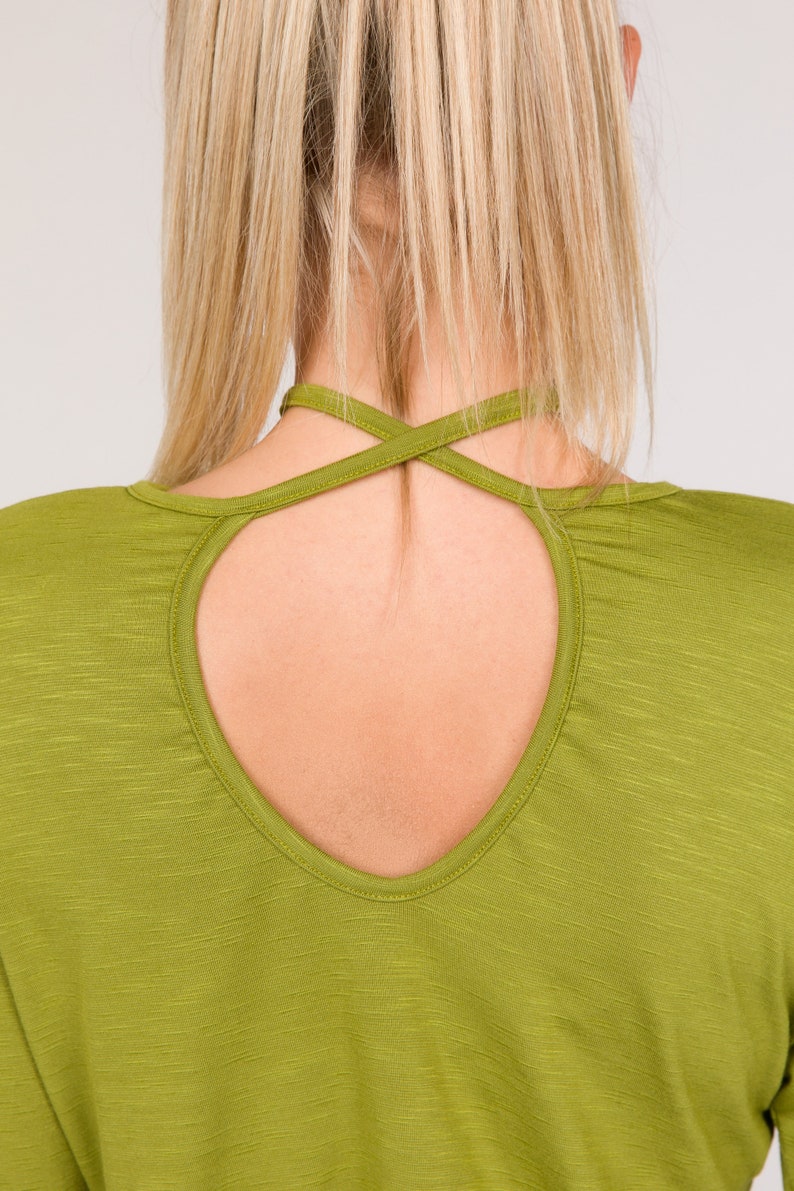 Puede incluir: Primer plano de una camisa verde de manga larga con un dise&ntilde;o de espalda con abertura. La camisa tiene un escote redondo y un detalle de correas cruzadas en la espalda. La tela tiene una sutil apariencia texturizada.