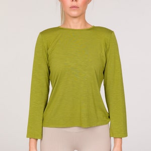Puede incluir: Una camiseta verde oliva de manga larga. La camiseta tiene cuello redondo y una silueta ajustada. La modelo tambi&eacute;n lleva leggings beiges. El fondo es de color neutro.