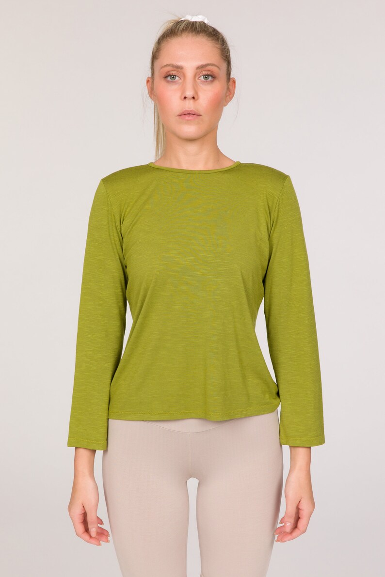 Puede incluir: Una camiseta verde oliva de manga larga. La camiseta tiene cuello redondo y una silueta ajustada. La modelo tambi&eacute;n lleva leggings beiges. El fondo es de color neutro.