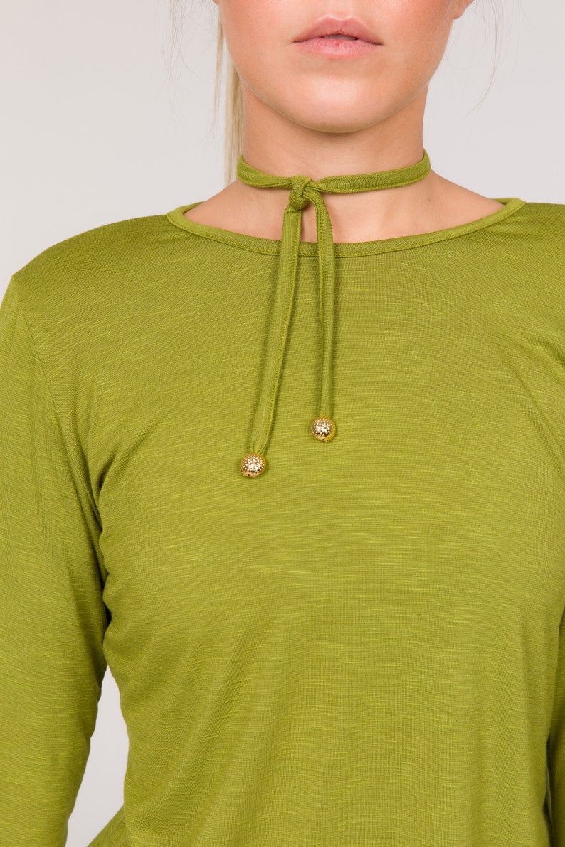 Puede incluir: Top verde oliva de manga larga con gargantilla a juego. La gargantilla tiene cierre de lazo y detalles decorativos de bolas doradas. El top tiene cuello redondo y un aspecto texturizado.