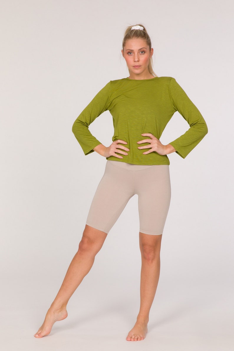 Puede incluir: Una persona con una camiseta verde oliva de manga larga y unos pantalones cortos de ciclista beiges. La camiseta tiene cuello redondo y los pantalones cortos llegan a la rodilla. El conjunto es sencillo y adecuado para actividades deportivas o informales.