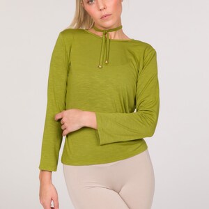 Puede incluir: Top verde oliva de manga larga con cuello redondo y cuello tipo gargantilla. El top tiene un detalle de cord&oacute;n en el escote con detalles dorados. Combinado con un pantal&oacute;n corto de ciclista beige, el conjunto est&aacute; sobre un fondo blanco.