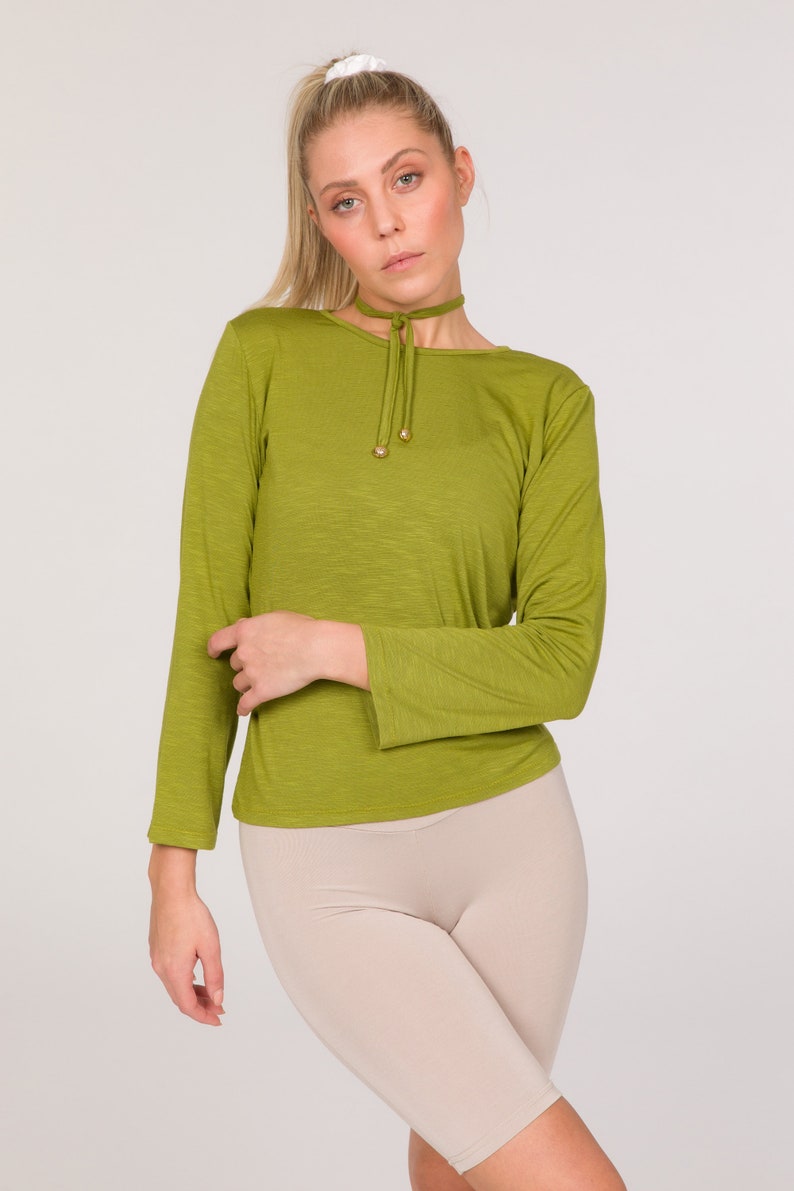Puede incluir: Top verde oliva de manga larga con cuello redondo y cuello tipo gargantilla. El top tiene un detalle de cord&oacute;n en el escote con detalles dorados. Combinado con un pantal&oacute;n corto de ciclista beige, el conjunto est&aacute; sobre un fondo blanco.