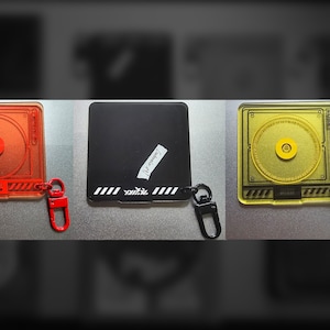 May include: Three acrylic keychains: red, black, and yellow. The red keychain is a square with a CD player design and a red clasp. The black keychain is square with a white stripe and text. The yellow keychain has a CD player design and a yellow clasp.