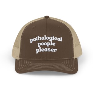 May include: A brown trucker hat with a tan mesh back. The hat features the white text "pathological people pleaser" on the front. The brim is brown, and the hat has a classic trucker style.