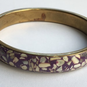 May include: A brass bangle bracelet with a purple and white speckled design. The bracelet is made of metal and has a smooth, polished finish.