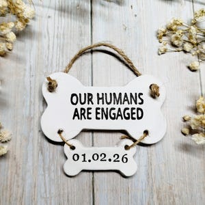 May include: A white, bone-shaped sign with the text "OUR HUMANS ARE ENGAGED" in black. Below, a smaller bone-shaped sign reads "01.02.26". The sign is suspended by a twine loop. Decorative flowers surround the sign.