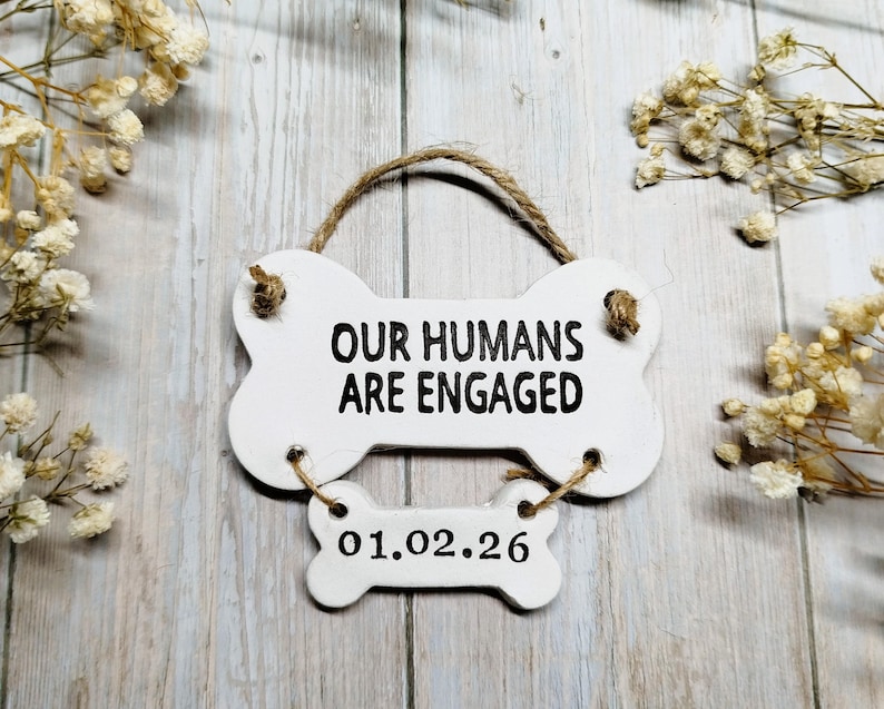 May include: A white, bone-shaped sign with the text "OUR HUMANS ARE ENGAGED" in black. Below, a smaller bone-shaped sign reads "01.02.26". The sign is suspended by a twine loop. Decorative flowers surround the sign.