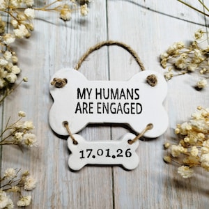 May include: A white, bone-shaped sign with the text "MY HUMANS ARE ENGAGED" and the date "17.01.26" in black. The sign is suspended by a twine loop and sits on a light wood surface, surrounded by small white flowers.
