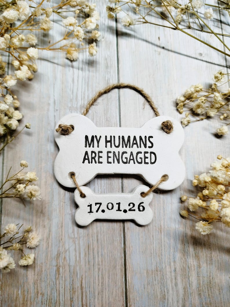 May include: A white, bone-shaped sign with the text "MY HUMANS ARE ENGAGED" and the date "17.01.26" in black. The sign is suspended by a twine loop and sits on a light wood surface, surrounded by small white flowers.