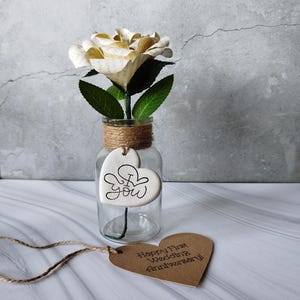 May include: A clear glass vase with a paper rose and green leaves, tied with twine. A heart-shaped tag with "I love you" is attached. A brown heart-shaped tag with "Happy First Wedding Anniversary" is also present.
