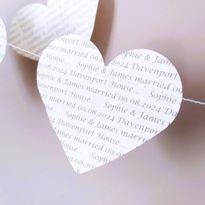 May include: A string of white paper heart decorations. Each heart is printed with the text "Sophie & James married 09.08.2024 Davenport House..." in a repeating pattern. The hearts are strung together with a thin white string.