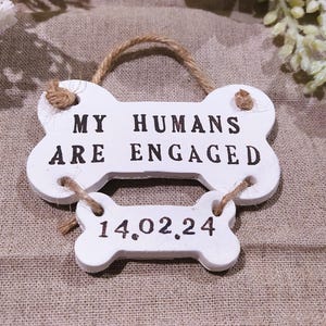 May include: White bone-shaped sign with the text "MY HUMANS ARE ENGAGED" and the date "14.02.24" in black. The sign is made of wood and has a twine hanger. The background is a neutral fabric.
