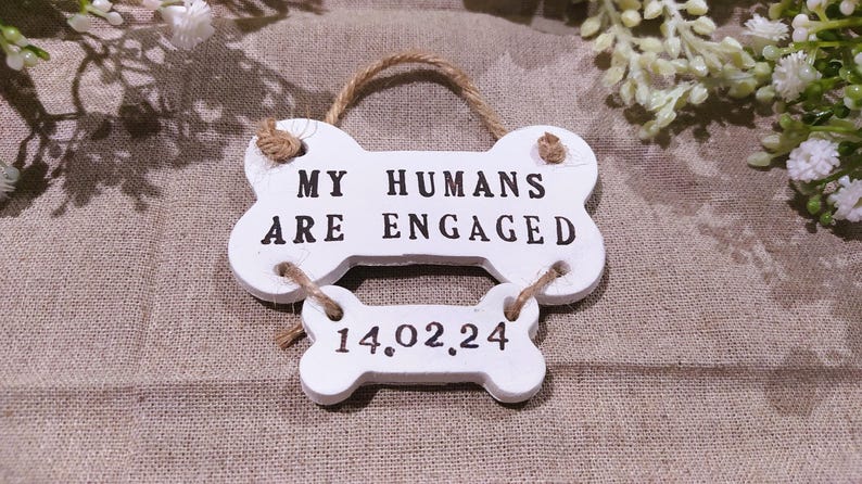 May include: White bone-shaped sign with the text "MY HUMANS ARE ENGAGED" and the date "14.02.24" in black. The sign is made of wood and has a twine hanger. The background is a neutral fabric.