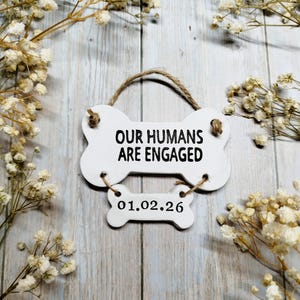 May include: A white, bone-shaped sign with the text "OUR HUMANS ARE ENGAGED" and the date "01.02.26". The sign is suspended by a twine loop and surrounded by dried white flowers.