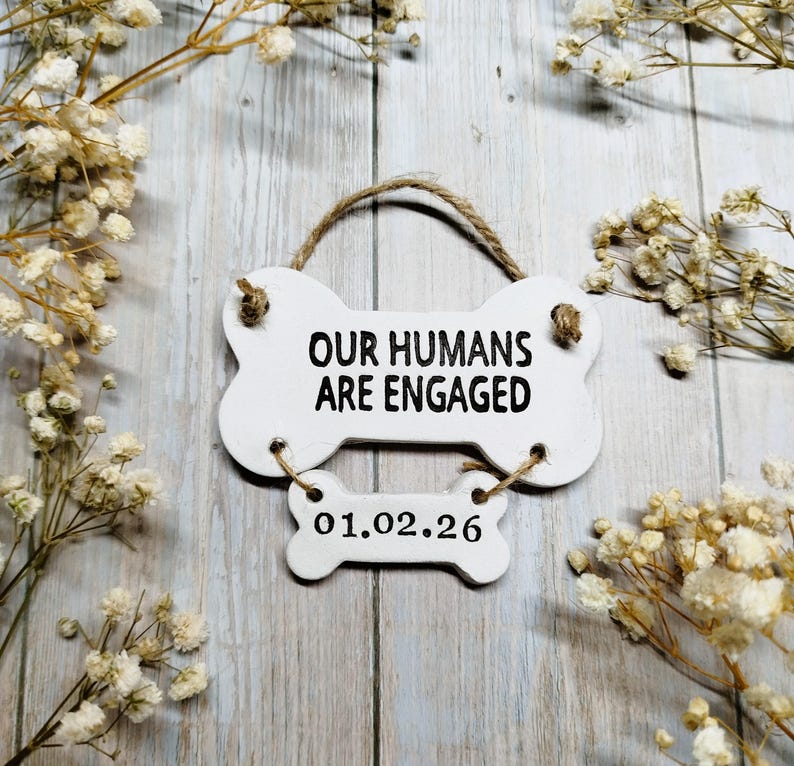 May include: A white, bone-shaped sign with the text "OUR HUMANS ARE ENGAGED" and the date "01.02.26". The sign is suspended by a twine loop and surrounded by dried white flowers.