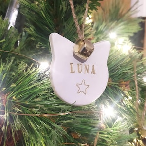 May include: A white cat-shaped Christmas decoration with the name "LUNA" in gold lettering and a gold star. A small gold bell hangs above the cat's head. The ornament is suspended by a natural twine string and is displayed on a Christmas tree.