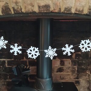 May include: A white snowflake garland strung across a fireplace. The garland features multiple white snowflake cutouts, each with intricate designs. The background includes a brick fireplace and a dark metal pipe.