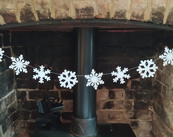 May include: A white snowflake garland strung across a fireplace. The garland features multiple white snowflake cutouts, each with intricate designs. The background includes a brick fireplace and a dark metal pipe.