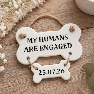 May include: Two white ceramic bone-shaped plaques connected by jute twine. The larger top bone features the black text MY HUMANS ARE ENGAGED, while the smaller bottom bone displays the date 25.07.26 in black font. The pieces hang from a twine loop.