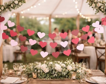 May include: A romantic outdoor wedding reception setup with heart-shaped garland in shades of red and pink. The table is decorated with white flowers, candles, and place settings. Soft lighting and a blurred background create a dreamy atmosphere.