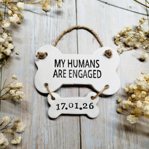 May include: A white, bone-shaped sign with the text "MY HUMANS ARE ENGAGED" in black. Below, a smaller bone-shaped sign displays the date "17.01.26". The sign is suspended by a twine loop and adorned with dried flowers.