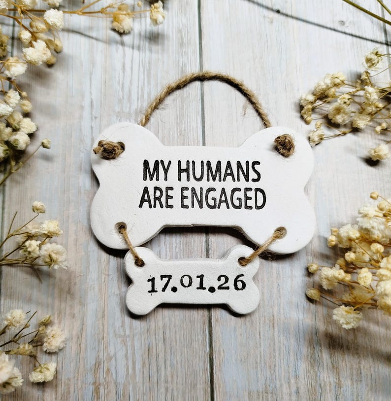 May include: A white, bone-shaped sign with the text "MY HUMANS ARE ENGAGED" in black. Below, a smaller bone-shaped sign displays the date "17.01.26". The sign is suspended by a twine loop and adorned with dried flowers.