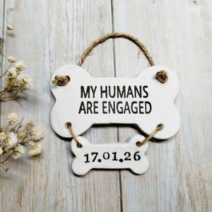 May include: A white, bone-shaped sign with the text "MY HUMANS ARE ENGAGED" in black lettering. Below, a smaller bone-shaped sign displays the date "17.01.26". The sign is suspended by a jute rope and is a charming engagement announcement.