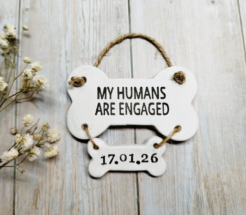 May include: A white, bone-shaped sign with the text "MY HUMANS ARE ENGAGED" in black lettering. Below, a smaller bone-shaped sign displays the date "17.01.26". The sign is suspended by a jute rope and is a charming engagement announcement.