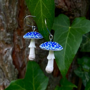 Op de afbeelding: Oorbellen in de vorm van paddenstoelen met blauwe doppen met witte stippen en witte stelen. Ze hangen voor groene bladeren en een boomstam.