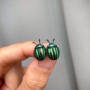 Op de afbeelding: Een paar oorknopjes in de vorm van kevers met metallic groene en zwarte gestreepte schilden. Elke kever heeft een rond lichaam, een gladde iriserende groene kop en twee kleine zwarte voelsprieten boven op de kop.