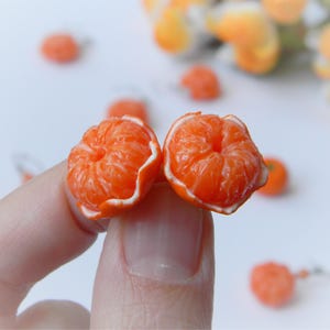 Könnte beinhalten: Ein Paar orangefarbene Ohrringe in Form von Miniatur-Mandarinen. Die Ohrringe werden zwischen Fingern gehalten, wodurch die detaillierte Textur und die weißen Ränder der Frucht hervorgehoben werden. Weitere orangefarbene Ohrringe sind im Hintergrund zu sehen.