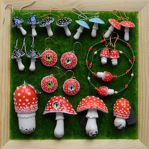 Op de afbeelding: Een verzameling handgemaakte paddenstoeloorbellen en een ketting. De oorbellen zijn gemaakt van polymer clay en hebben verschillende ontwerpen, waaronder rood-witte gestippelde paddenstoelen, zwart-witte gestippelde paddenstoelen, blauwe paddenstoelen en paddenstoelen met rode ogen. De ketting is gemaakt van groene en rode kralen en heeft een rood-witte gestippelde paddenstoelbedel.