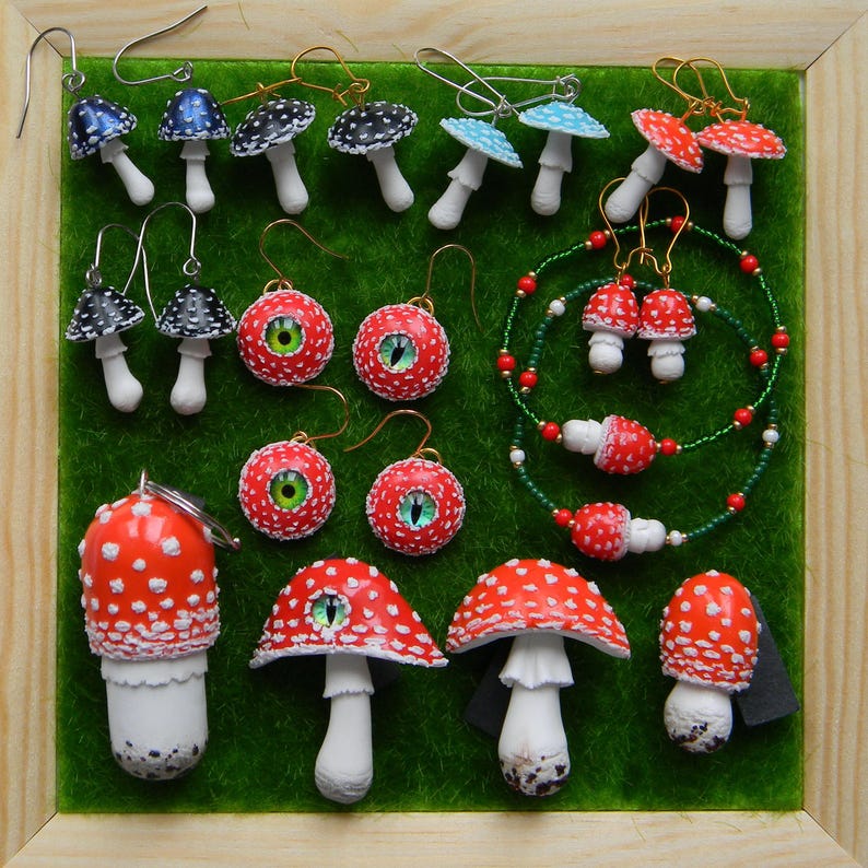 Op de afbeelding: Een verzameling handgemaakte paddenstoeloorbellen en een ketting. De oorbellen zijn gemaakt van polymer clay en hebben verschillende ontwerpen, waaronder rood-witte gestippelde paddenstoelen, zwart-witte gestippelde paddenstoelen, blauwe paddenstoelen en paddenstoelen met rode ogen. De ketting is gemaakt van groene en rode kralen en heeft een rood-witte gestippelde paddenstoelbedel.