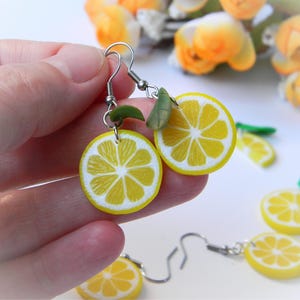 Könnte beinhalten: Handgefertigte Ohrringe mit Zitronenscheiben. Jeder Ohrring hat ein gelb-weißes Zitronenscheiben-Design mit einem grünen Blattakzent. Die Ohrringe werden in einer Hand gehalten, im Hintergrund sind weitere Ohrringe mit Zitronenscheiben zu sehen.